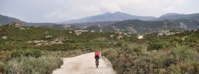 Cycling. Offroad Cycling in the Agriates Desert. Corsica, France. a red shirt cyclist, Dusty road, Rocky road, Mediterranean Vegetation. Mountains, Daylight. Cloudy sky. Courage for the Journey.
