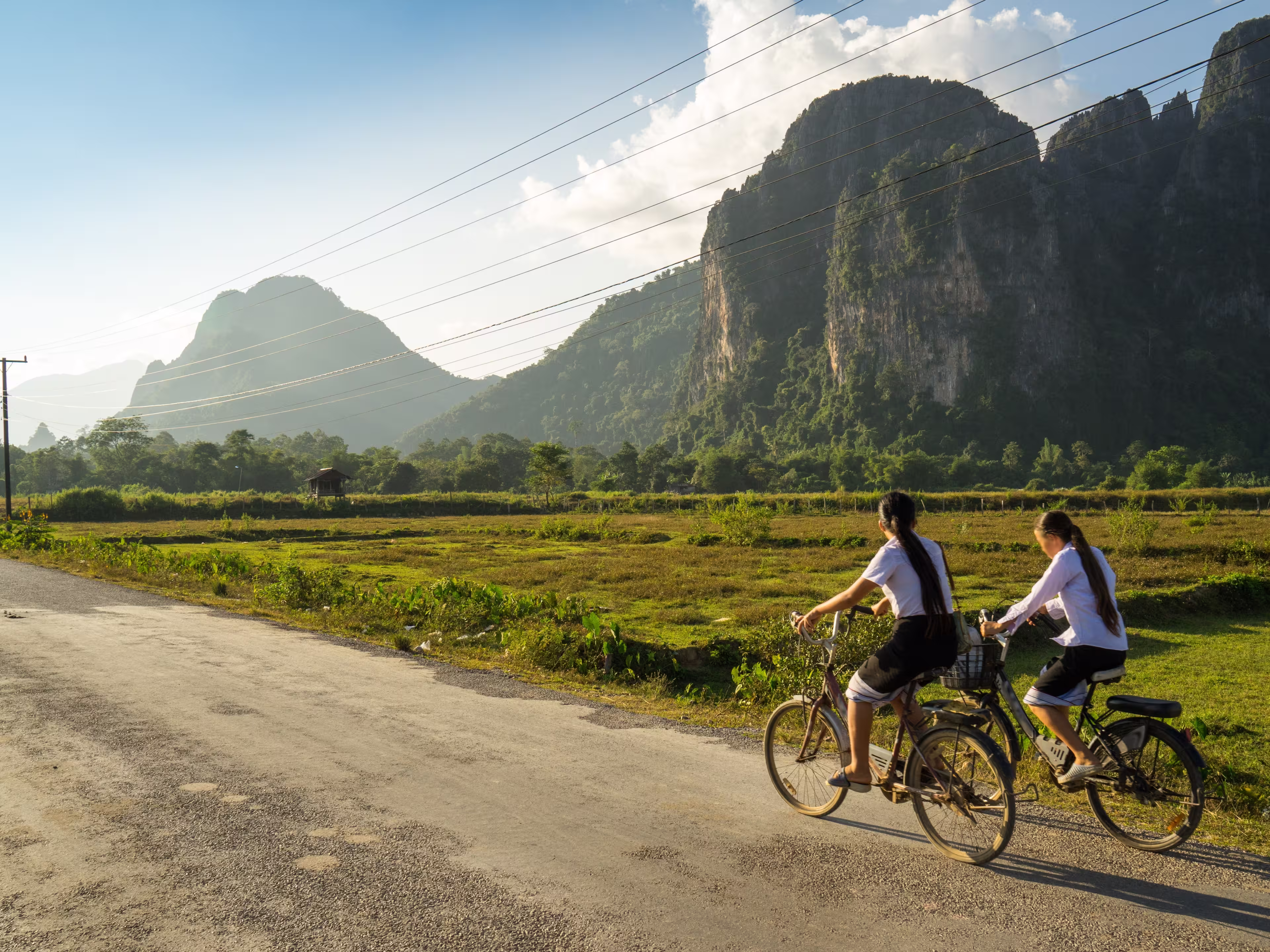 Two schoolgirls cycle through the countryside of Vang Vieng, Laos, Southeast Asia, on a sunny late afternoon, past typical Laotian karst landscapes