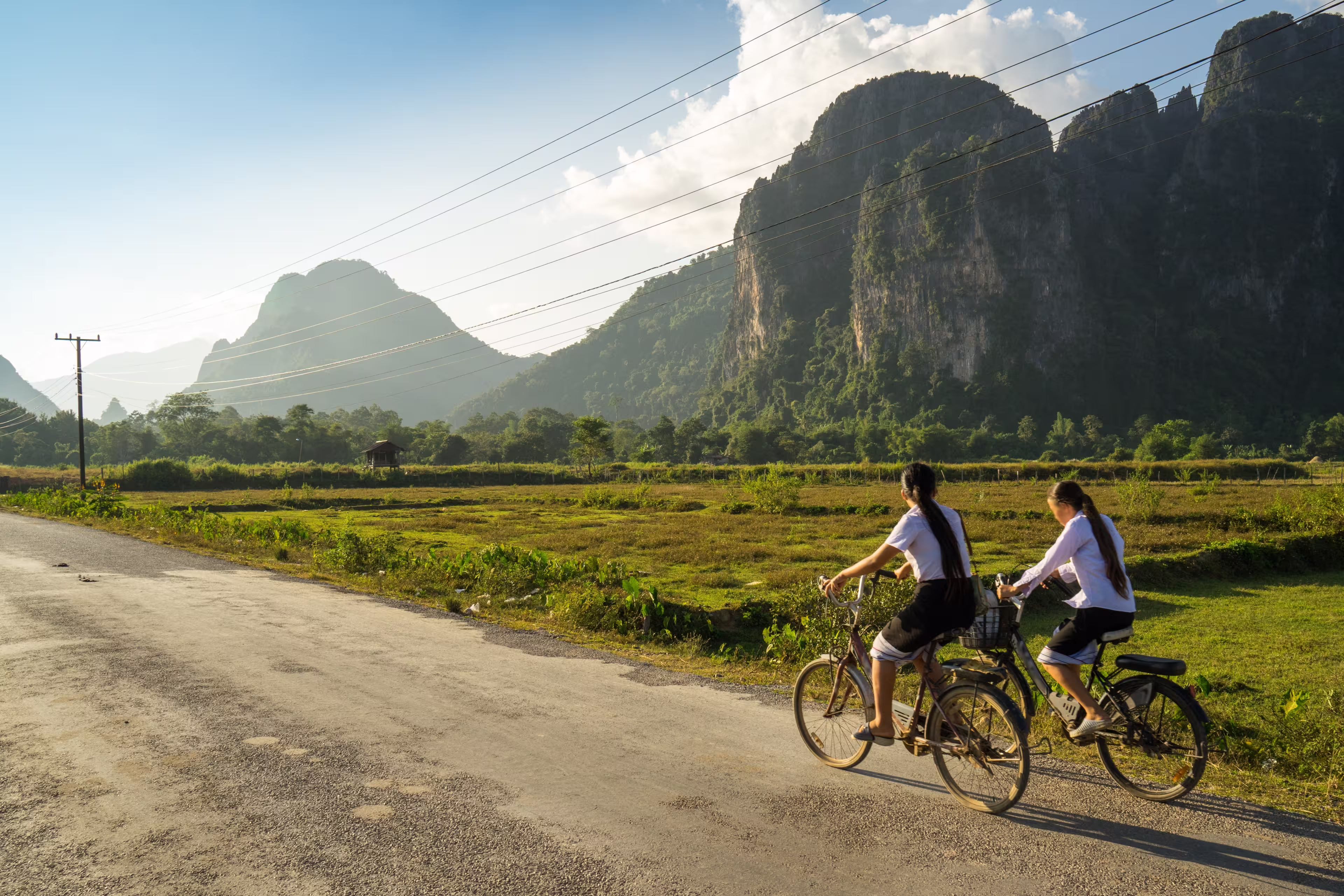 Two schoolgirls cycle through the countryside of Vang Vieng, Laos, Southeast Asia, on a sunny late afternoon, past typical Laotian karst landscapes