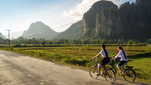 Two schoolgirls cycle through the countryside of Vang Vieng, Laos, Southeast Asia, on a sunny late afternoon, past typical Laotian karst landscapes