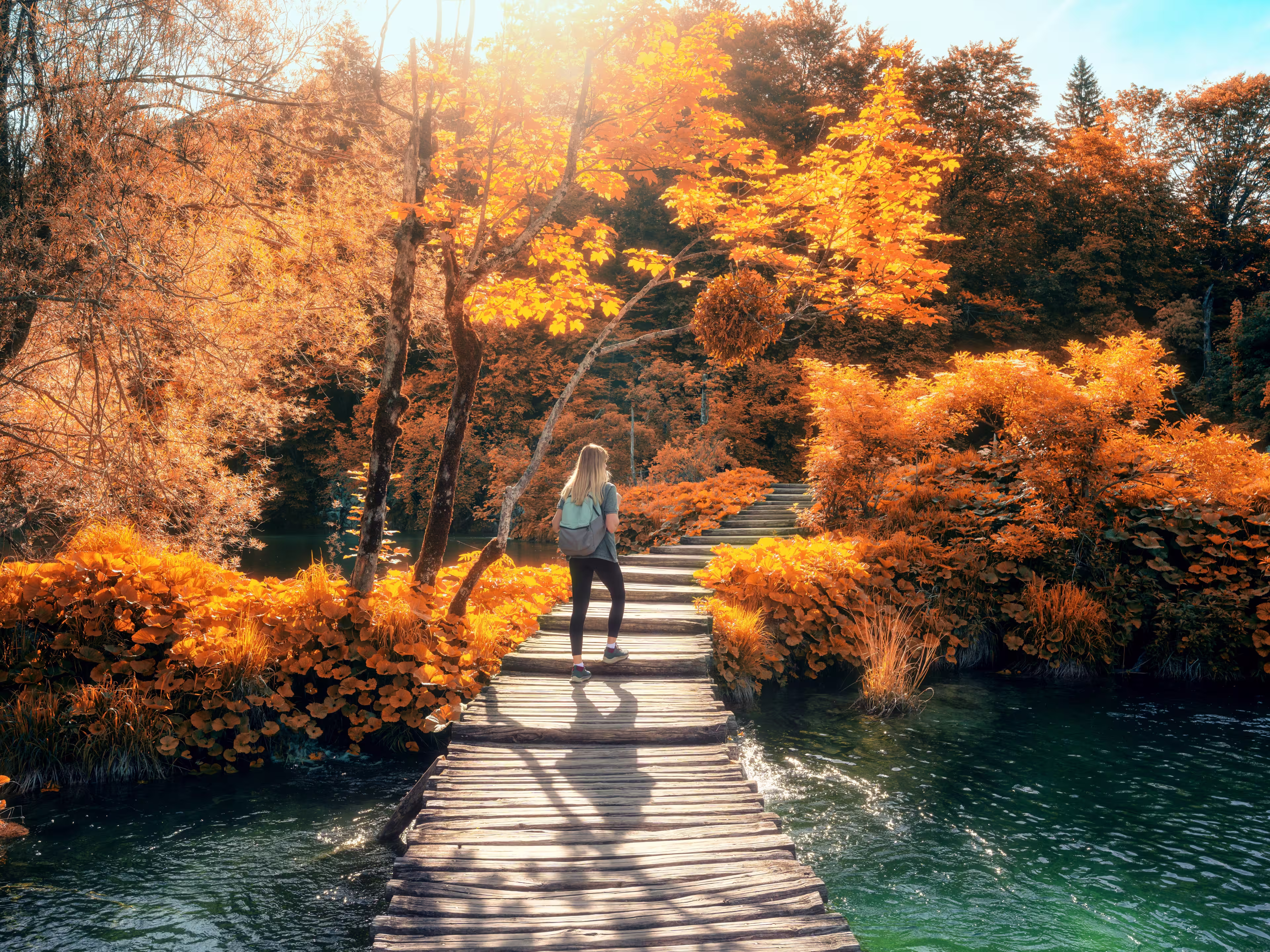 Young woman with backpack walking along a wooden path near stunning waterfalls, orange trees in Plitvice Lakes national park, Croatia at sunset in autumn. Sporty girl in forest in fall. Hiking, travel