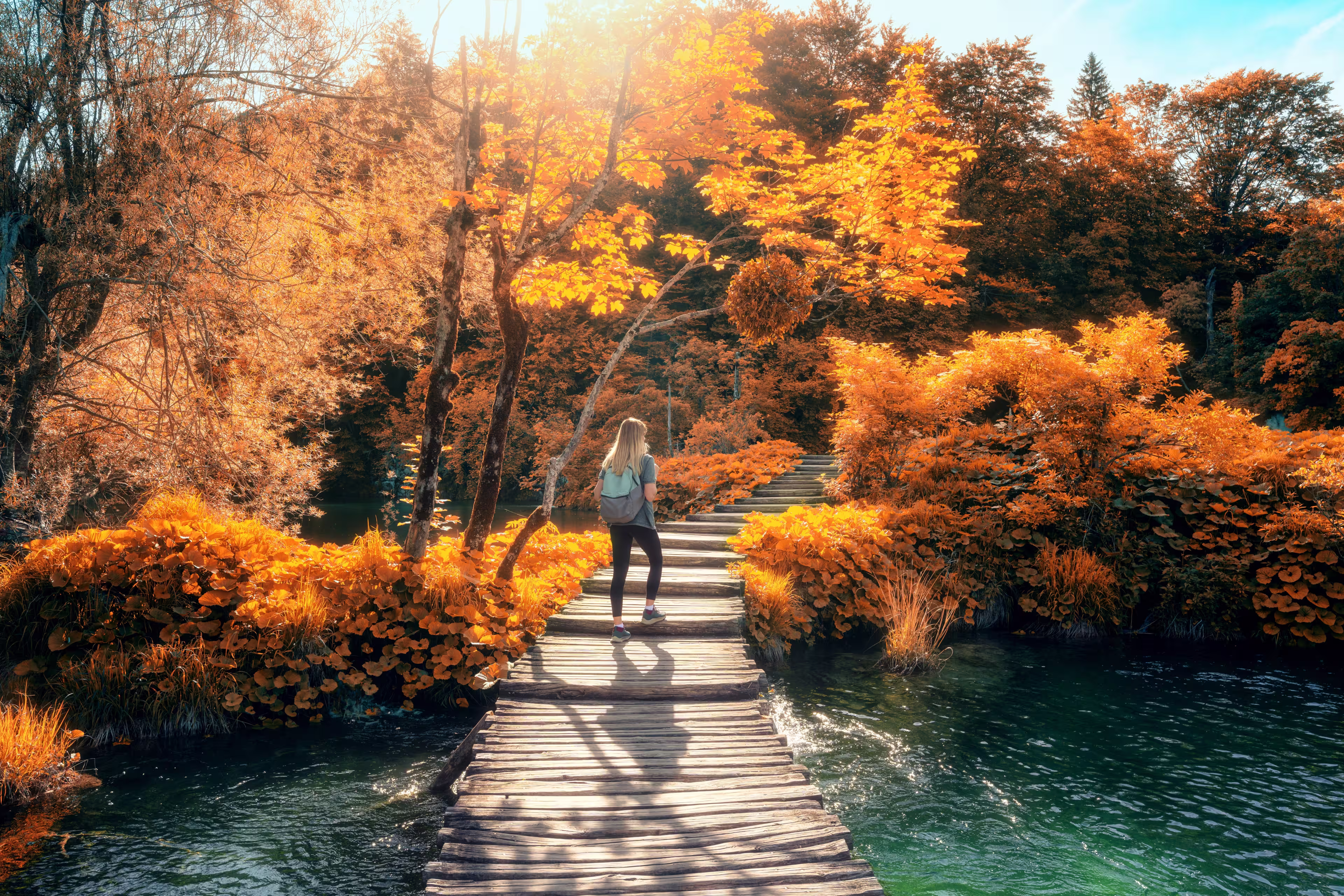 Young woman with backpack walking along a wooden path near stunning waterfalls, orange trees in Plitvice Lakes national park, Croatia at sunset in autumn. Sporty girl in forest in fall. Hiking, travel