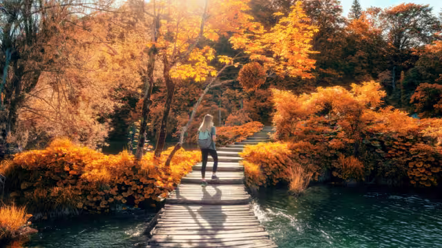 Young woman with backpack walking along a wooden path near stunning waterfalls, orange trees in Plitvice Lakes national park, Croatia at sunset in autumn. Sporty girl in forest in fall. Hiking, travel