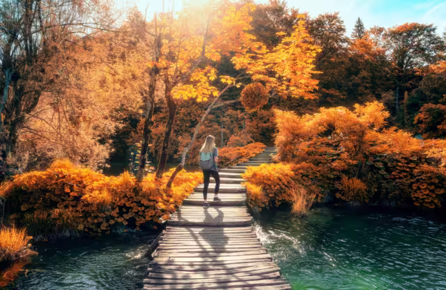 Young woman with backpack walking along a wooden path near stunning waterfalls, orange trees in Plitvice Lakes national park, Croatia at sunset in autumn. Sporty girl in forest in fall. Hiking, travel