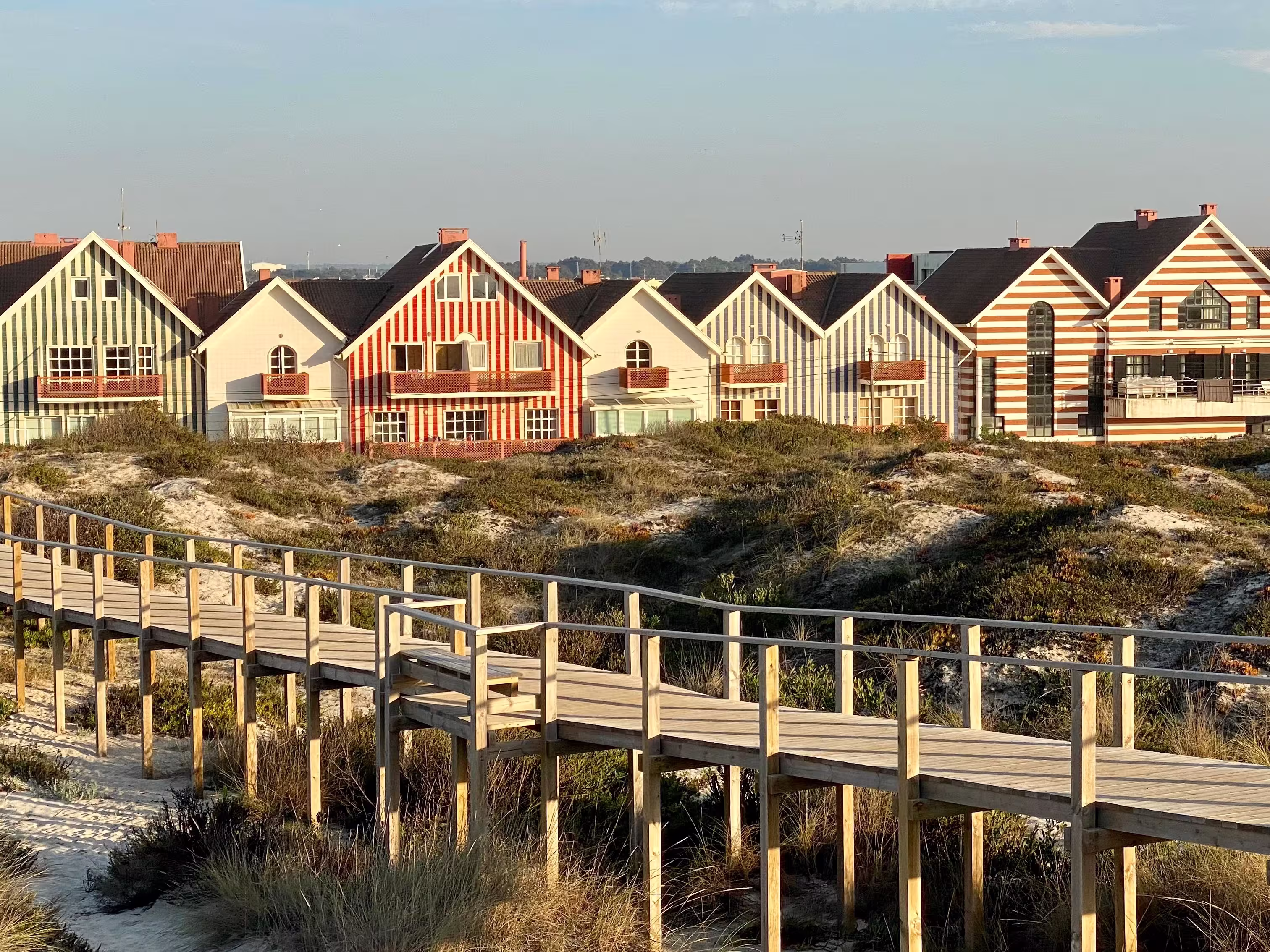 Striped coastal houses in Costa Nova, Portugal, with wooden boardwalk and sand dunes under bright summer sky — traditional seaside architecture by the Atlantic Ocean