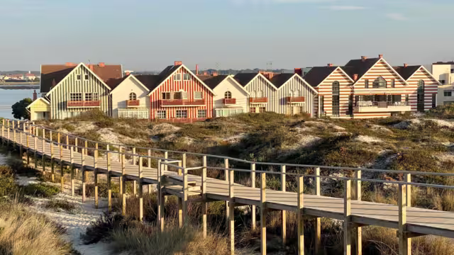 Striped coastal houses in Costa Nova, Portugal, with wooden boardwalk and sand dunes under bright summer sky — traditional seaside architecture by the Atlantic Ocean