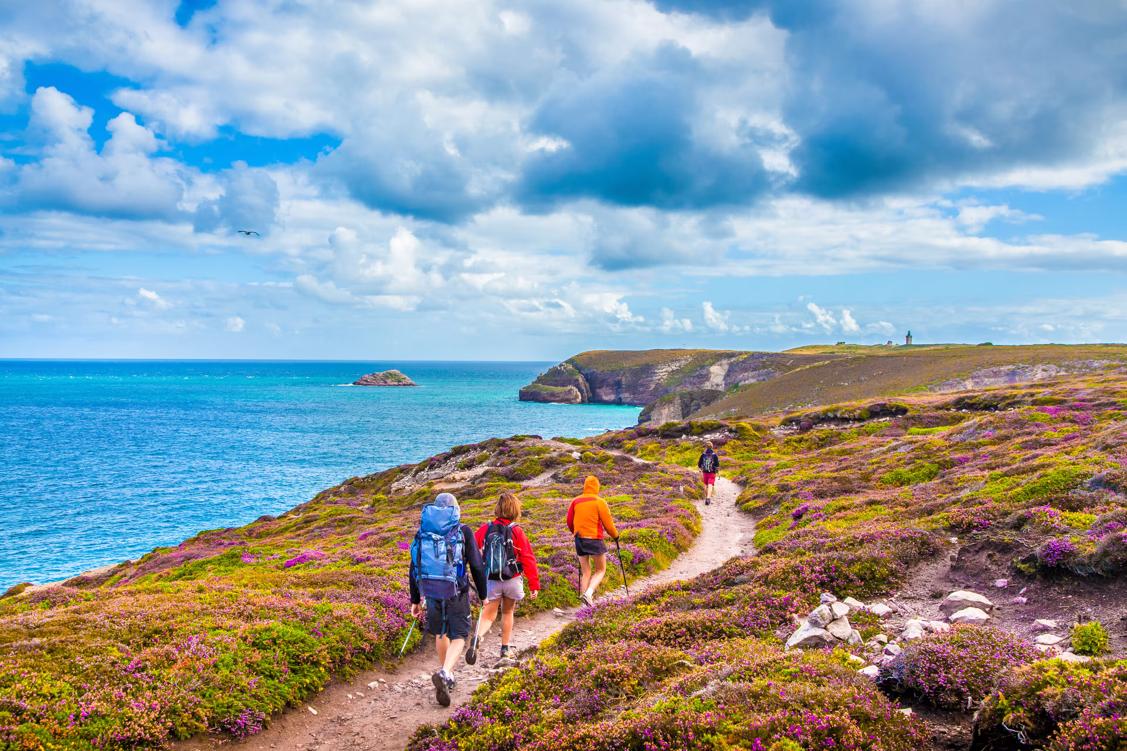 Tourists hiking along the beautiful coasts of Bretagne at famous Cap Frehel peninsula viewpoint with traditional lighthouse in the background on a sunny day with blue sky and clouds in summer, France