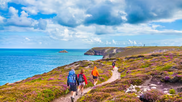 Tourists hiking along the beautiful coasts of Bretagne at famous Cap Frehel peninsula viewpoint with traditional lighthouse in the background on a sunny day with blue sky and clouds in summer, France