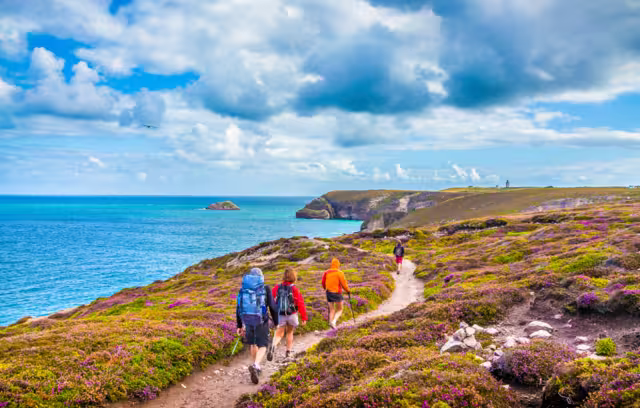Tourists hiking along the beautiful coasts of Bretagne at famous Cap Frehel peninsula viewpoint with traditional lighthouse in the background on a sunny day with blue sky and clouds in summer, France