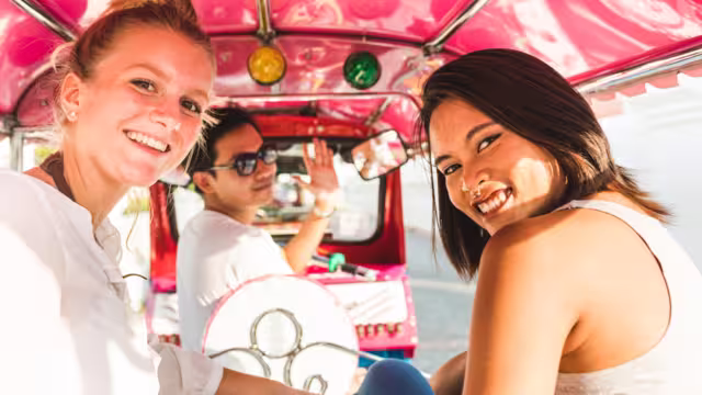 Friends smile at camera enjoying a tuk tuk ride in downtown Bangkok in Thailand