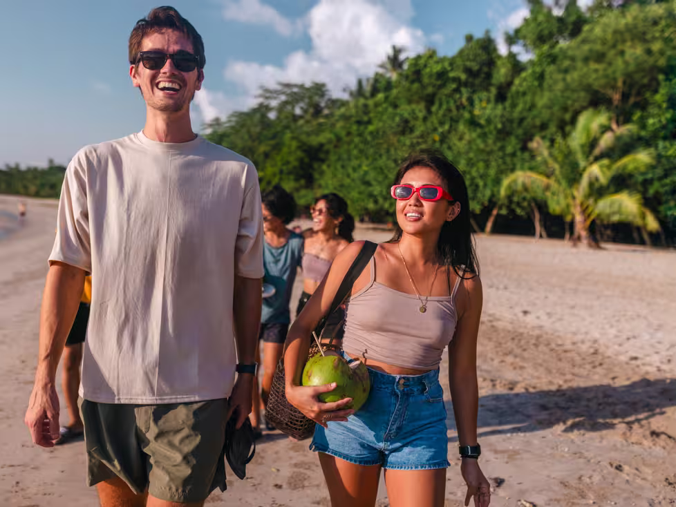 Young Intrepid travellers stroll along a beach in the Philippines with drinking coconuts laughing and enjoyng the sun